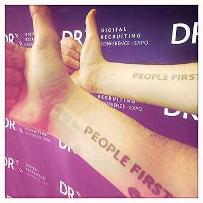 Zwei Unterarme mit Einwegtatoo 'People First' auf der Digital Recruiting Conference x Exposition 2024 vor Bannerhintergrund