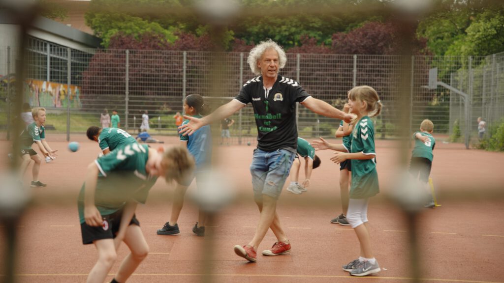 Ein Erzieher trainiert eine Gruppe von Kindern im Handball auf einem Handballplatz. Standbild aus der Episode 'Schule der besonderen Menschen' aus der Recruiting Video Serie für 'Die Bildungspartner' der Bewegtbildagentur DAS GUTE WERK.