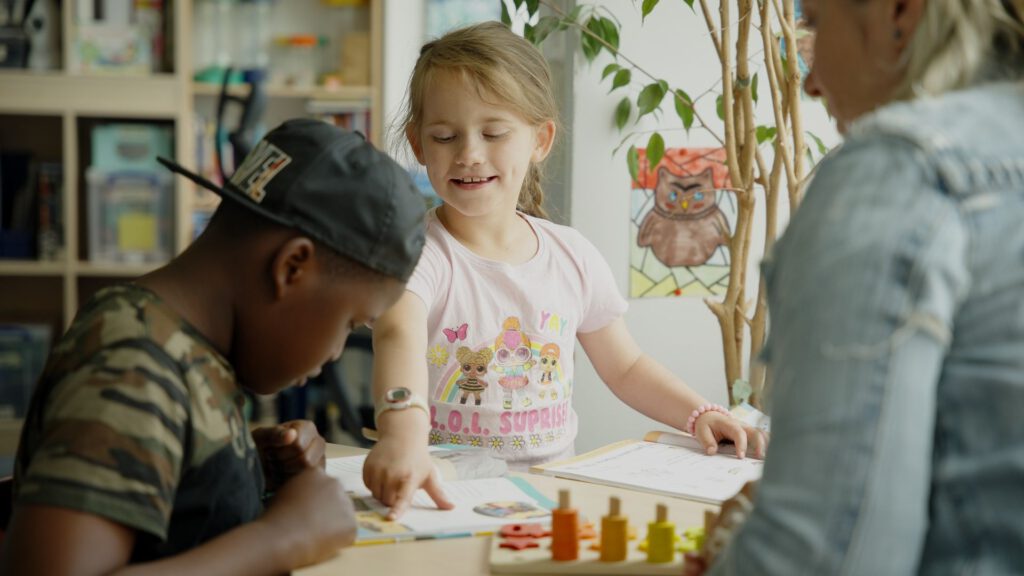 Ein Mädchen zeigt einem Jungen etwas in seinem Heft, eine Erzieherin schaut zu. Standbild aus der Recruiting Video Serie für 'Die Bildungspartner'.
